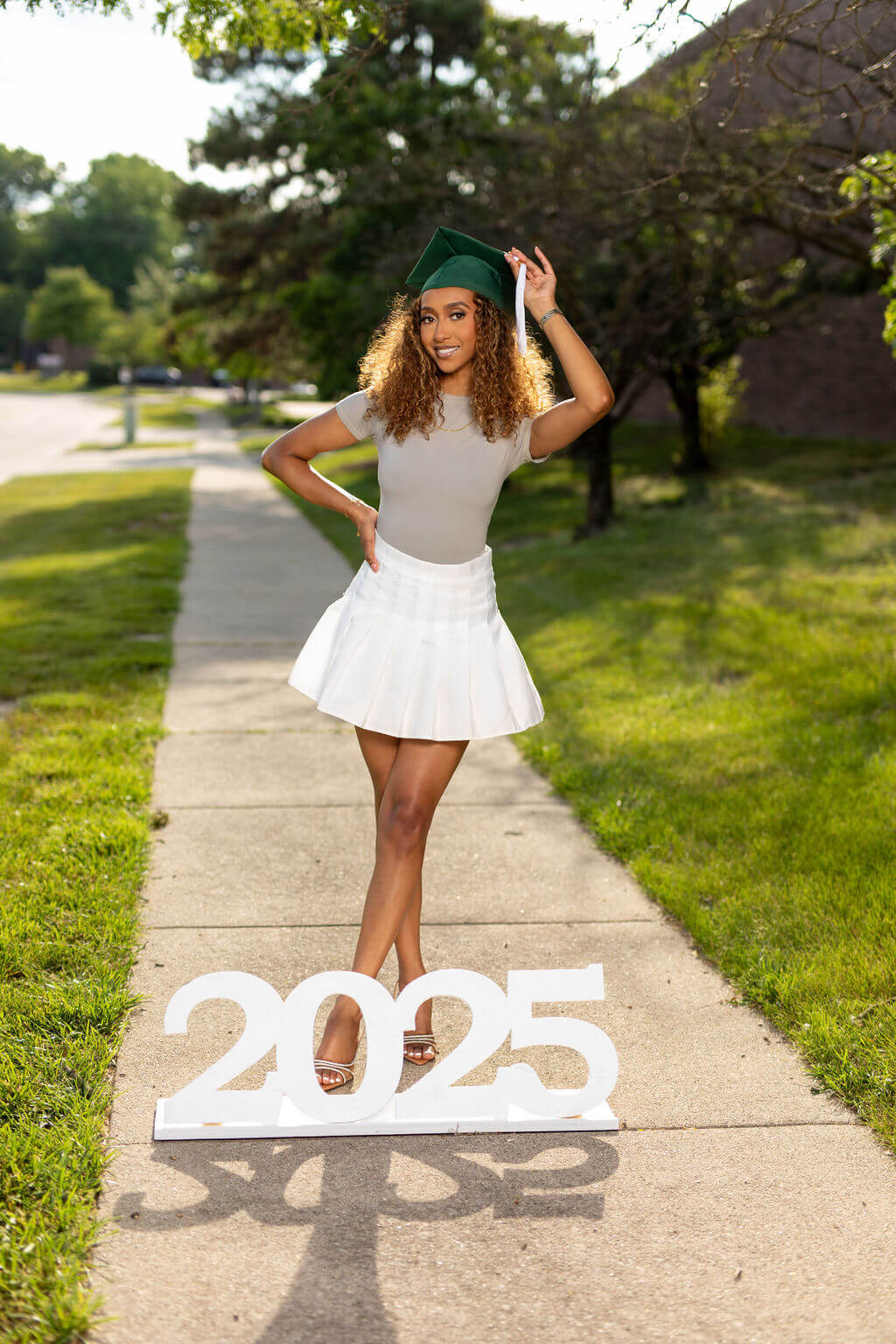 Senior Model standing on a sidewalk, holding her tassel, standing behind a giant "2025" photo prop!
