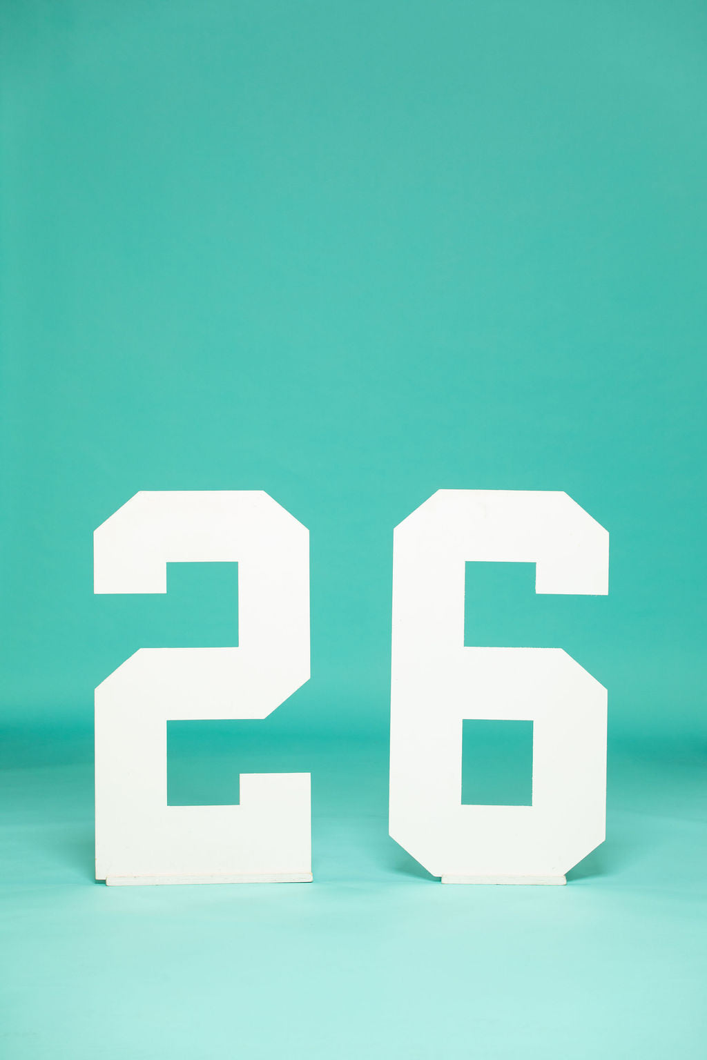 Bold white number props “26” on a seamless teal background, designed for class of 2026 senior pictures. Shop now at collageandwood.com.