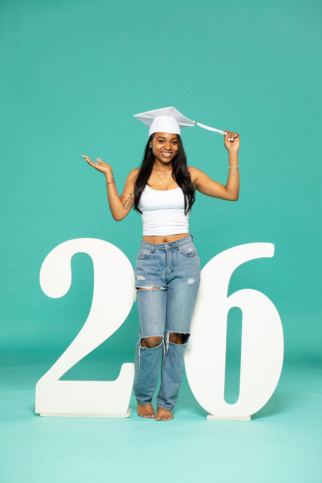 Female graduate in white cap and distressed jeans posing confidently between large script-style “26” numbers, holding her tassel. 2026 senior props available now at collageandwood.com.