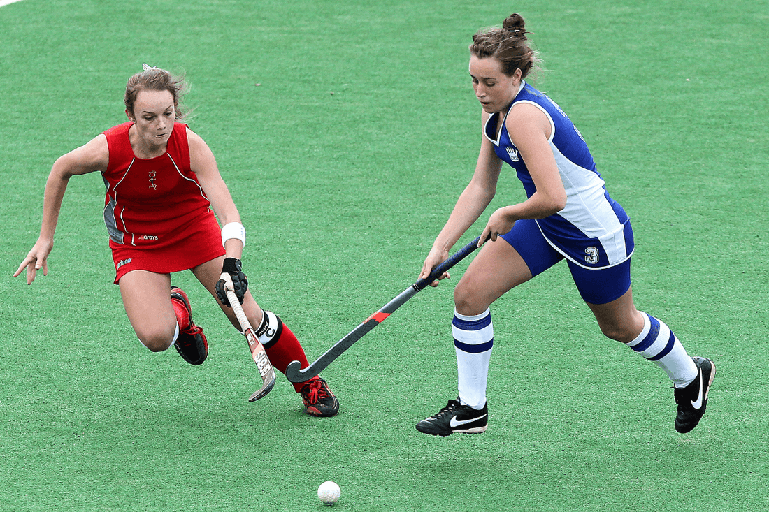 Two women competing in a field hockey game; one player in a red uniform lunges toward the ball while another in a blue and white uniform controls it with her stick on a green field.