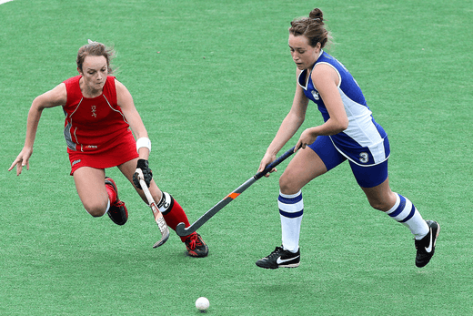 Two women competing in a field hockey game; one player in a red uniform lunges toward the ball while another in a blue and white uniform controls it with her stick on a green field. PHOTO BY CANVA