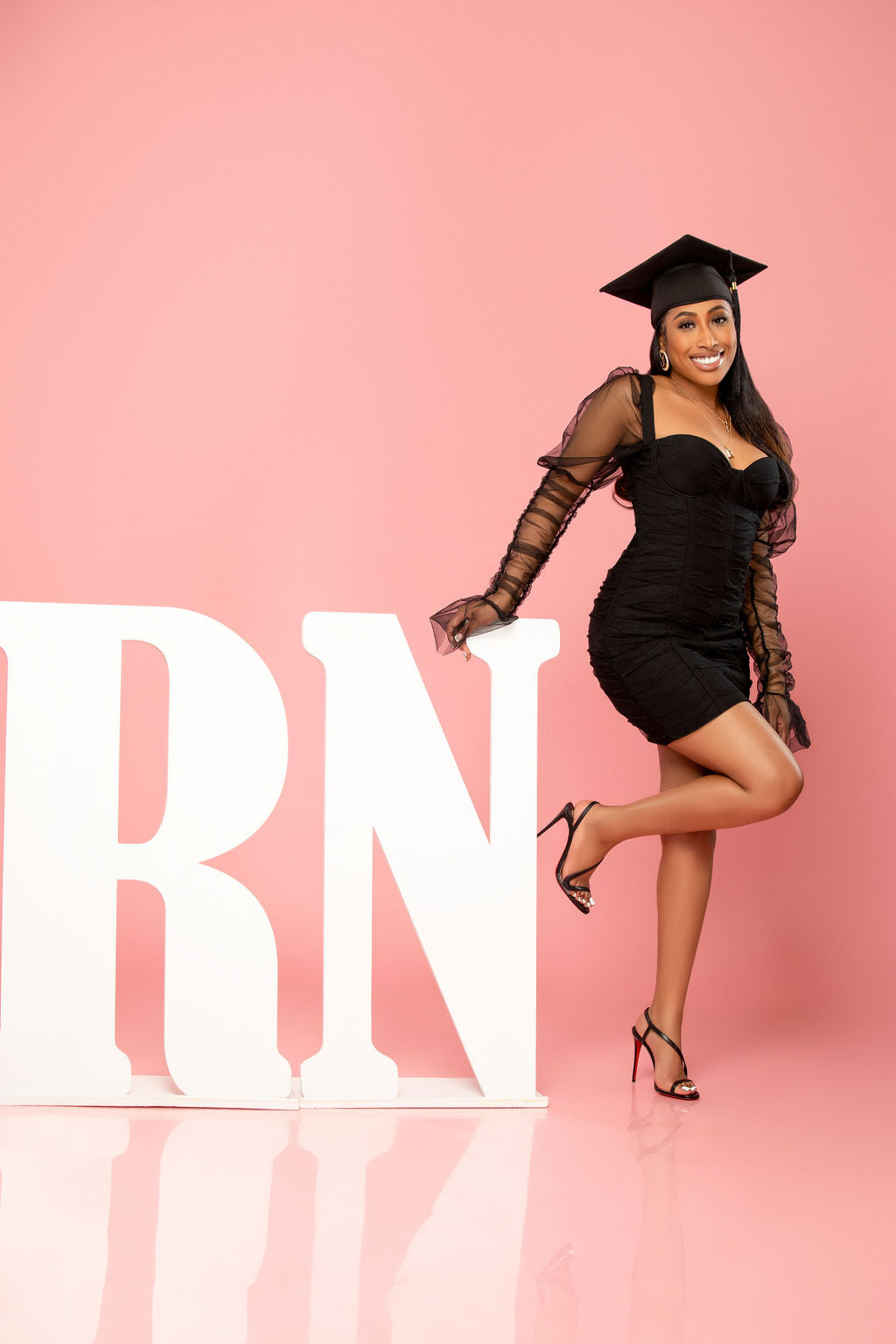 A woman wearing a stylish black dress and a graduation cap poses joyfully next to large white "RN" letters. She balances on one foot against a bright pink background, celebrating her achievement.