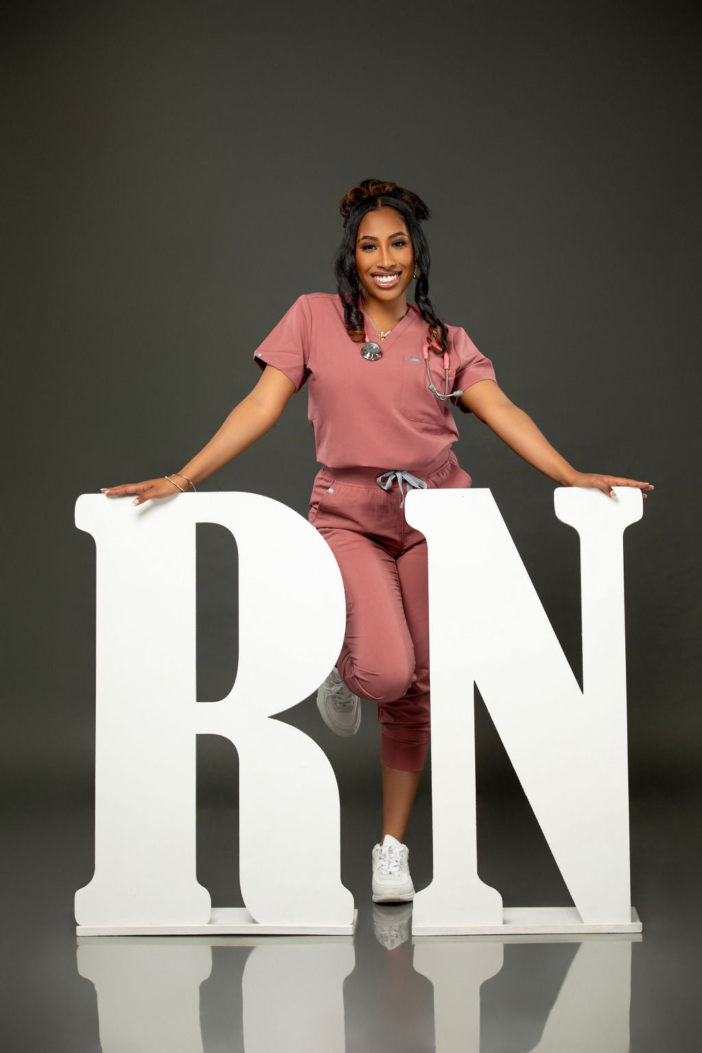 A woman wearing pink scrubs poses joyfully next to large white "RN" letters. She balances on one foot against a dark gray background, celebrating her achievement.