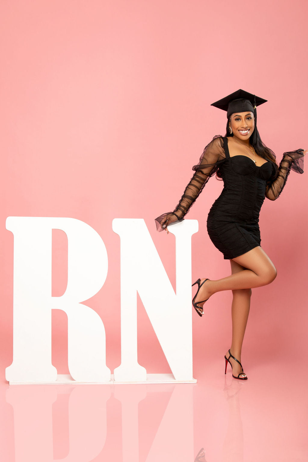 A woman wearing a stylish black dress and a graduation cap poses joyfully next to large white "RN" letters. She balances on one foot against a bright pink background, celebrating her achievement.