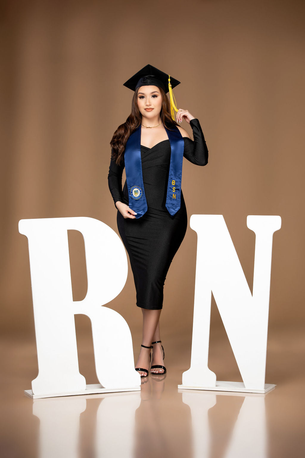 A woman wearing a stylish black dress and a graduation cap poses joyfully next to large white "RN" letters. She balances on one foot against a tan, celebrating her achievement.