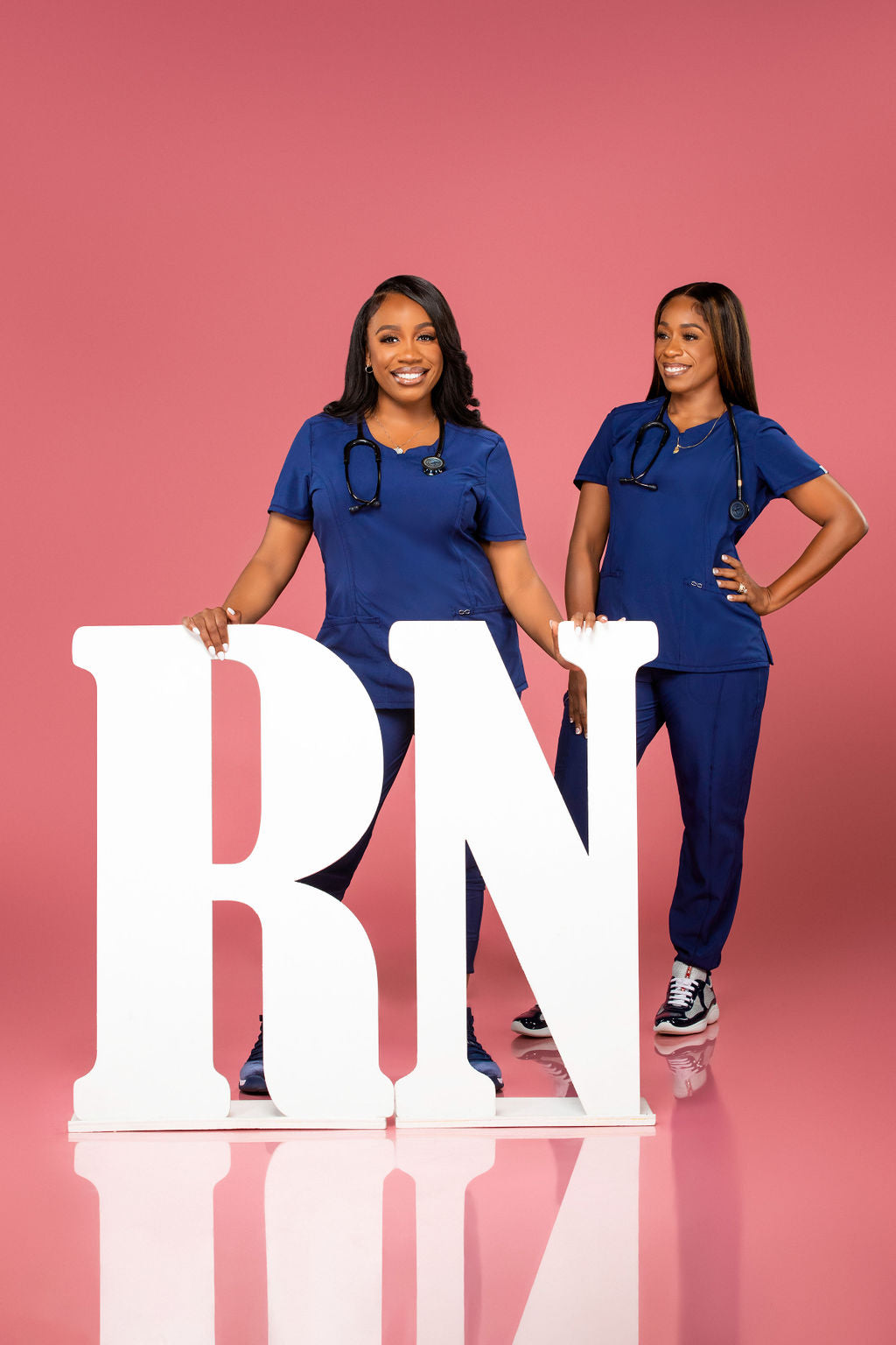 Two women in blue scrubs pose confidently next to large white "RN" letters against a bright pink background, celebrating their achievement.