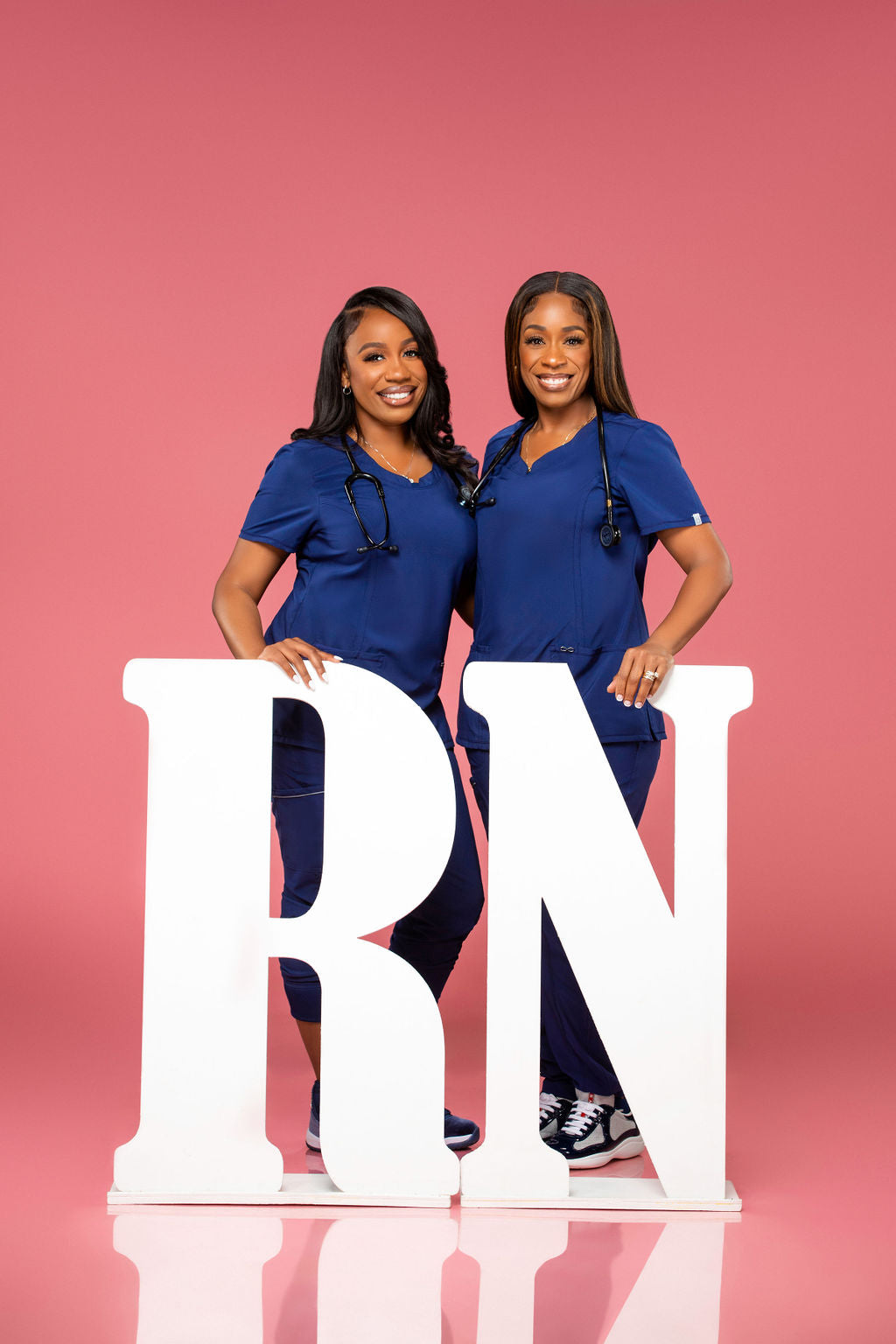 Two women in blue scrubs pose confidently next to large white "RN" letters against a bright pink background, celebrating their achievement.