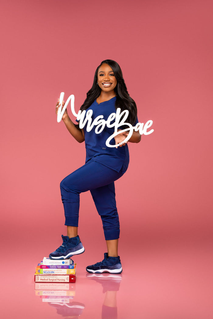 A woman wearing blue scrubs poses with "Nurse Bae" letters. She balances on one foot against a bright pink background, celebrating her achievement. Available at collageandwood.com.