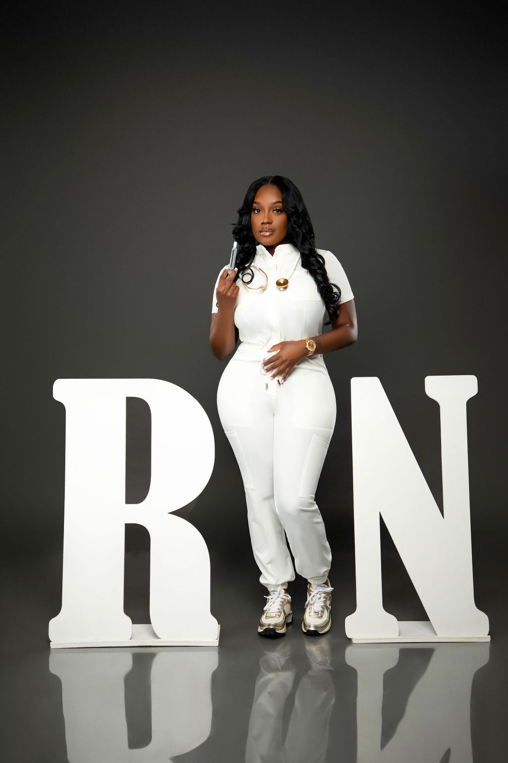 A young woman wearing pink scrubs with a stethoscope smiles confidently while leaning against large white letters spelling "RN." She stands on a reflective gray floor with a neutral dark gray background.