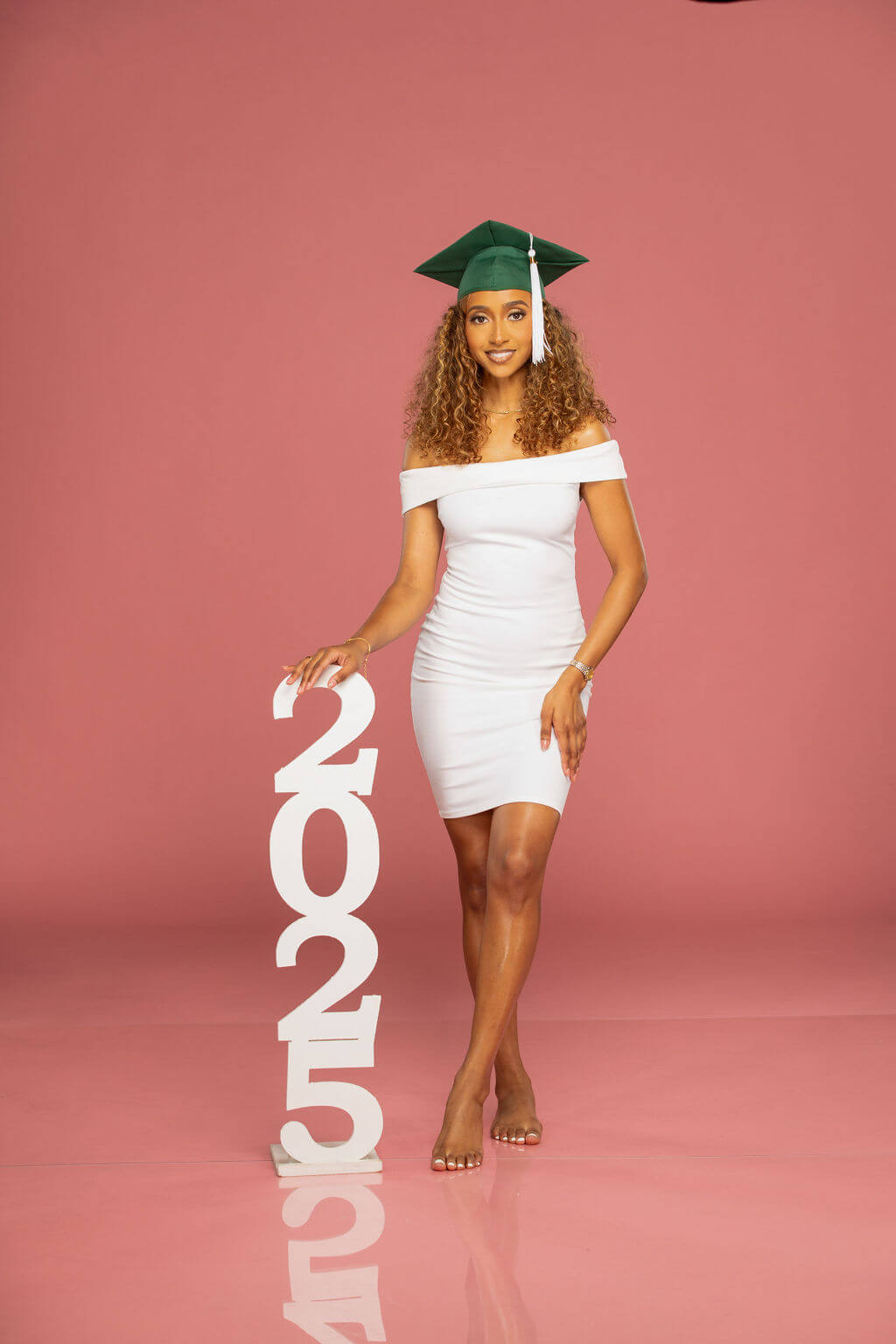 senior model in green graduation cap standing next to a 2025 photo vertical prop with a pink background.