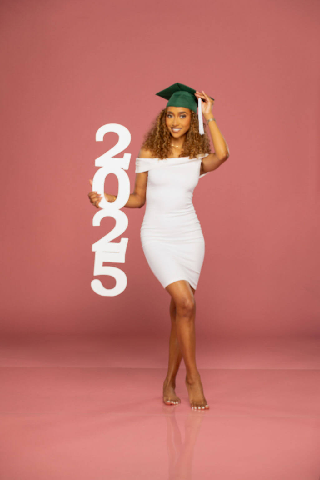 senior model in green graduation cap standing holding 2025 photo vertical prop with a pink background.