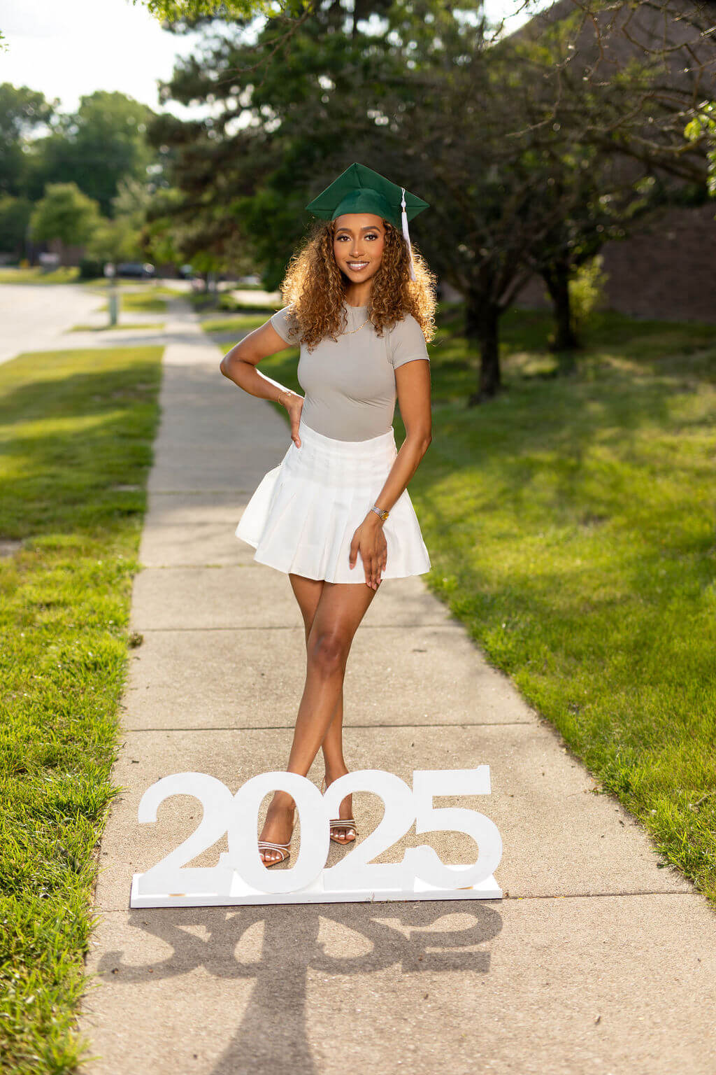 Senior Model standing on a sidewalk, holding her tassel, standing behind a giant "2025" photo prop!