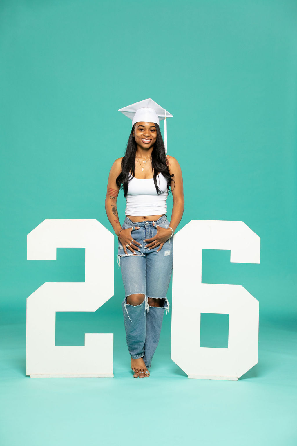 Female graduate in white cap and tank top standing between large block-style white numbers "2" and "6" on a teal background. 2026 senior props available now at collageandwood.com.