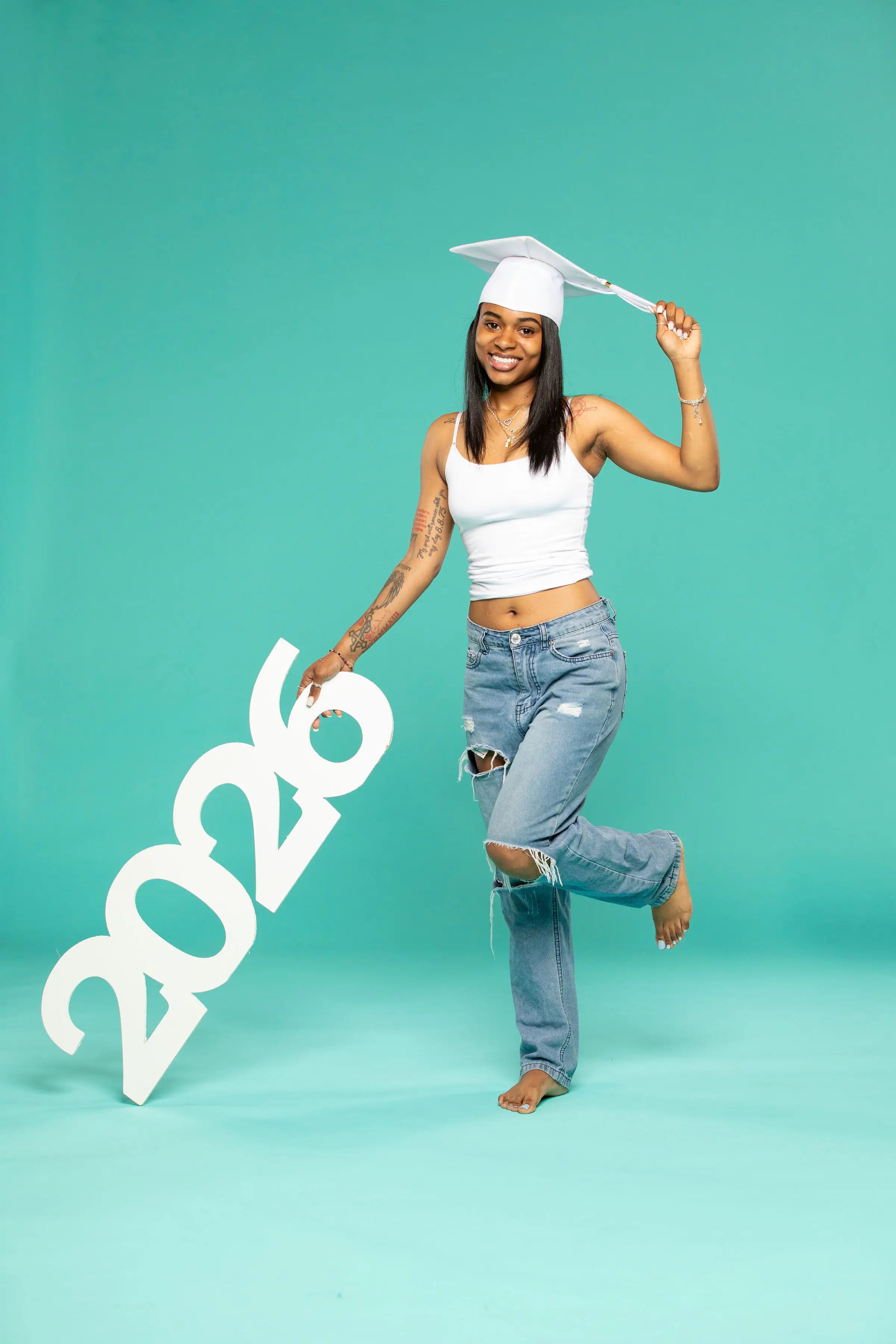 A smiling young woman wearing a white graduation cap and tank top holds a large white ‘2026’ prop from CollageandWood.com in front of her. She is barefoot, dressed in distressed jeans, and stands against a solid teal background."