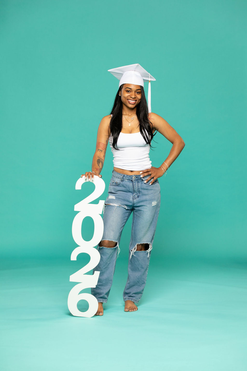 Female graduate in white cap and jeans stands confidently next to a vertical “2026” number prop on a teal background. 2026 senior props available now at collageandwood.com.