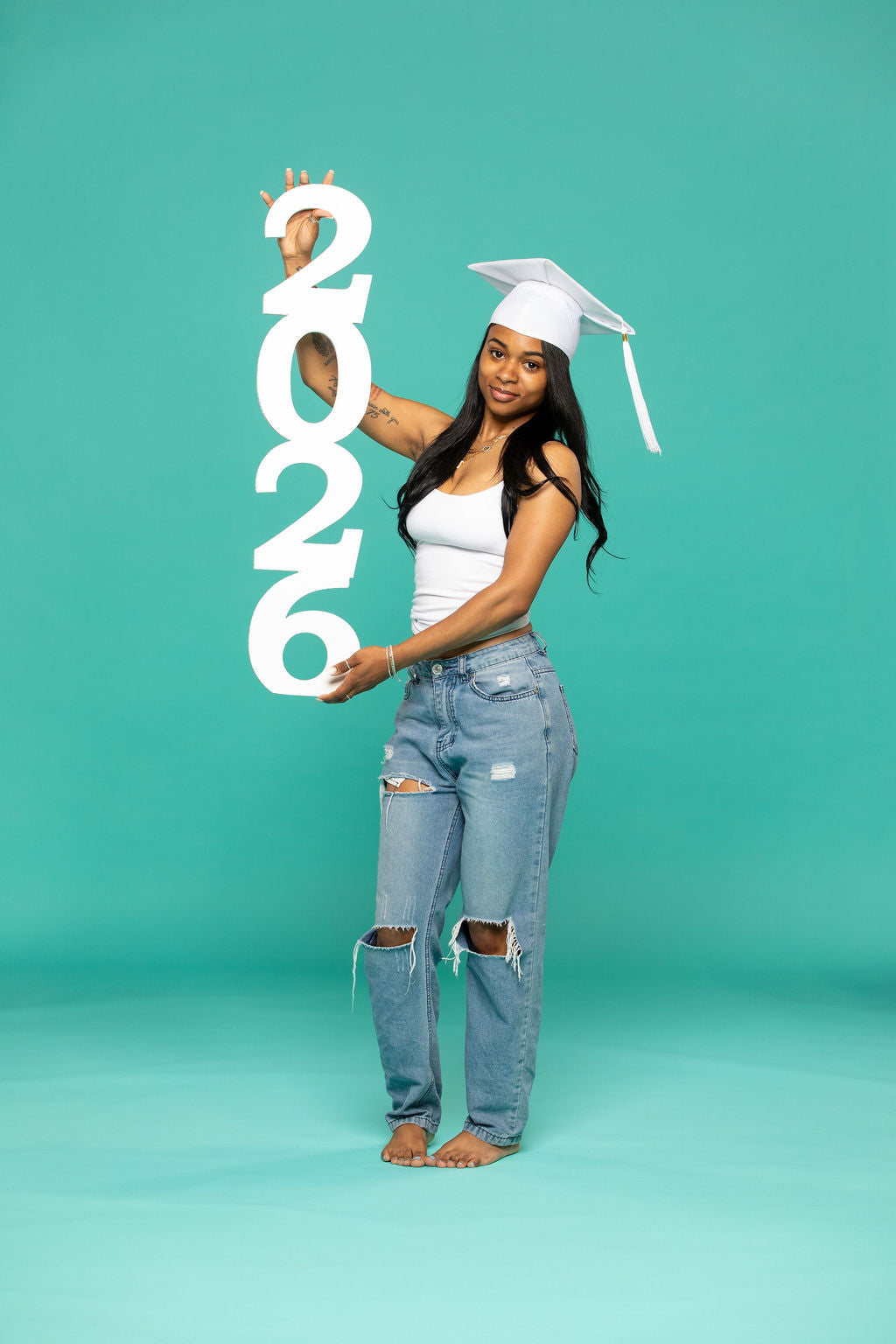 Class of 2026 graduate holding a tall “2026” number stack prop with both hands against a teal studio backdrop. 2026 senior props available now at collageandwood.com.