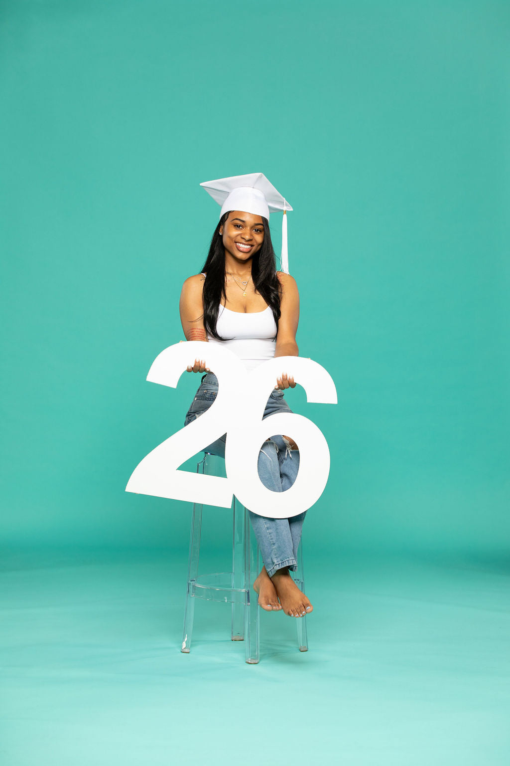 Young woman in a graduation cap sitting barefoot on a clear stool, holding a large white number "26" against a teal background. 2026 senior props available now at collageandwood.com.