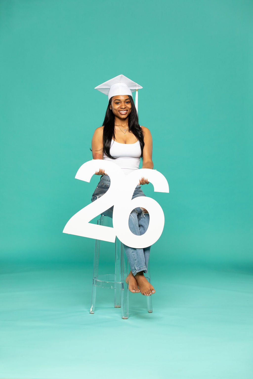 Woman in white graduation cap and distressed jeans sitting confidently on a clear stool, holding a white "26" in her lap. Props for 2026 seniors available at collageandwood.com.