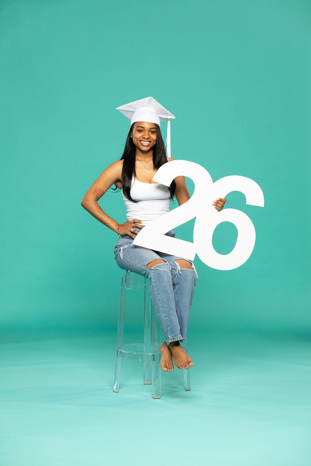 Smiling graduate in white cap and tank top sitting on a stool, displaying a bold white "26" in front of her. Celebrate with 2026 senior props from collageandwood.com.