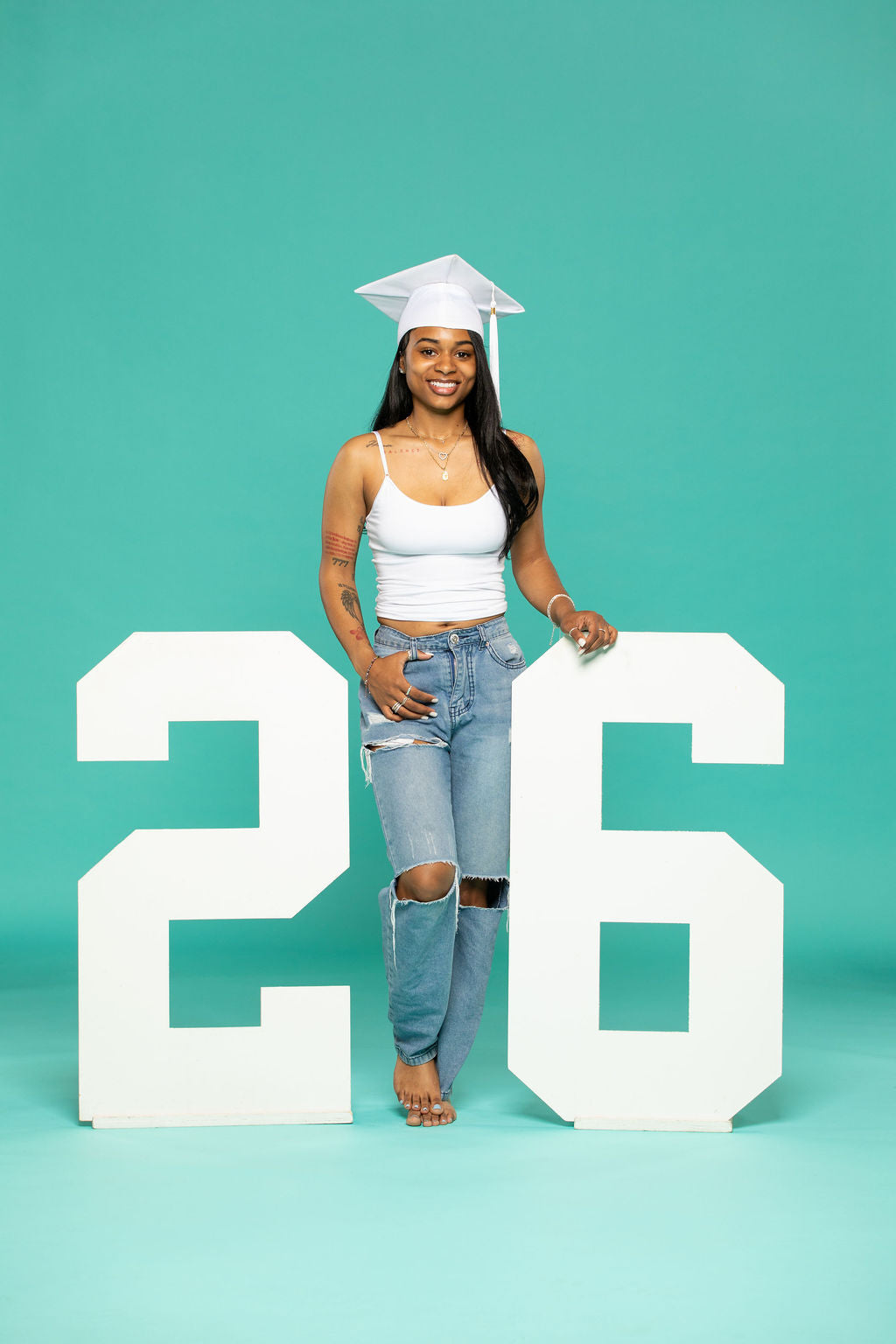 Smiling 2026 graduate in a white cap standing beside large white “26” numbers, perfect for senior photo shoots. Find props now at collageandwood.com.