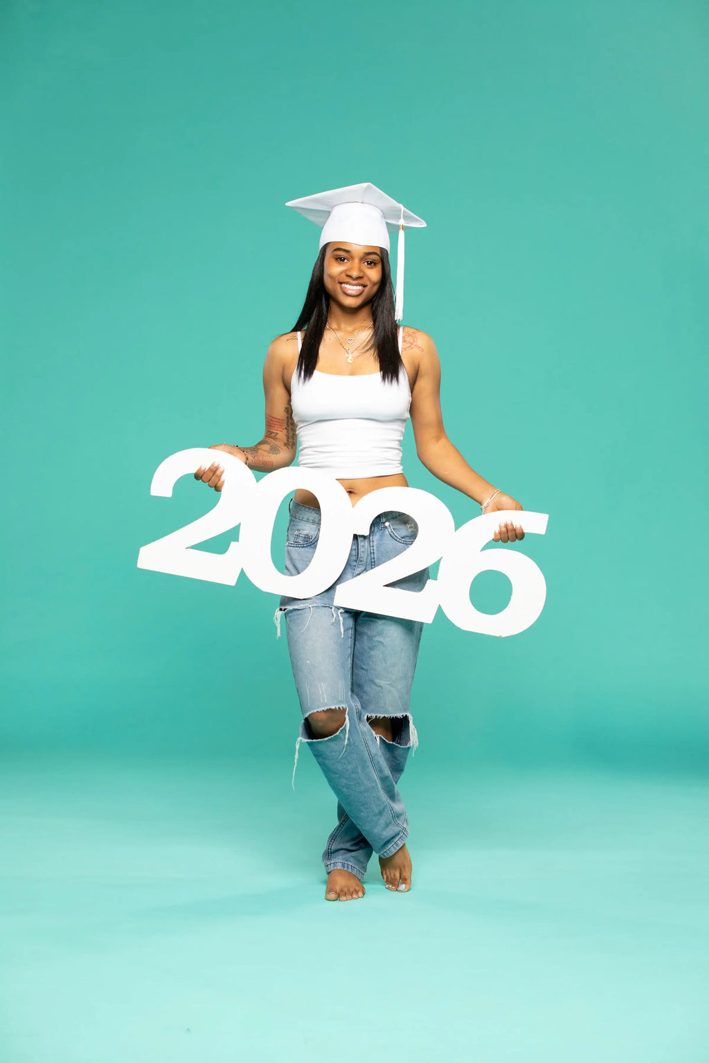 A barefoot young woman in a white graduation cap and tank top poses joyfully on one leg while holding a large white ‘2026’ photo prop from CollageandWood.com. She stands against a teal studio background, wearing ripped jeans and smiling confidently.