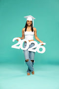 A barefoot young woman in a white graduation cap and tank top poses joyfully on one leg while holding a large white ‘2026’ photo prop from CollageandWood.com. She stands against a teal studio background, wearing ripped jeans and smiling confidently.