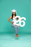 Female graduate in white cap and jeans poses barefoot on a stool with a large "26" number, teal backdrop. Shop 2026 senior photo props now at collageandwood.com.