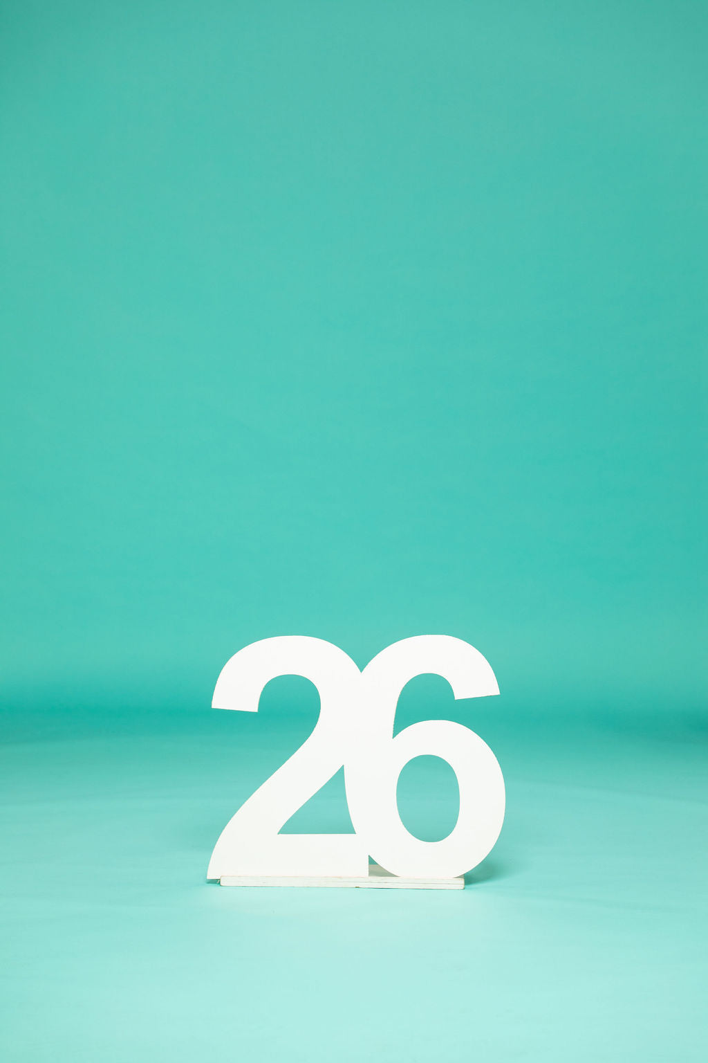 Large white number "26" standing alone on a teal studio floor with no people in frame. Perfect for 2026 senior photos—available now at collageandwood.com.