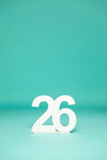 Minimalist shot of a white “26” number prop centered on a teal background, symbolizing a graduating class. Get your 2026 senior props at collageandwood.com.