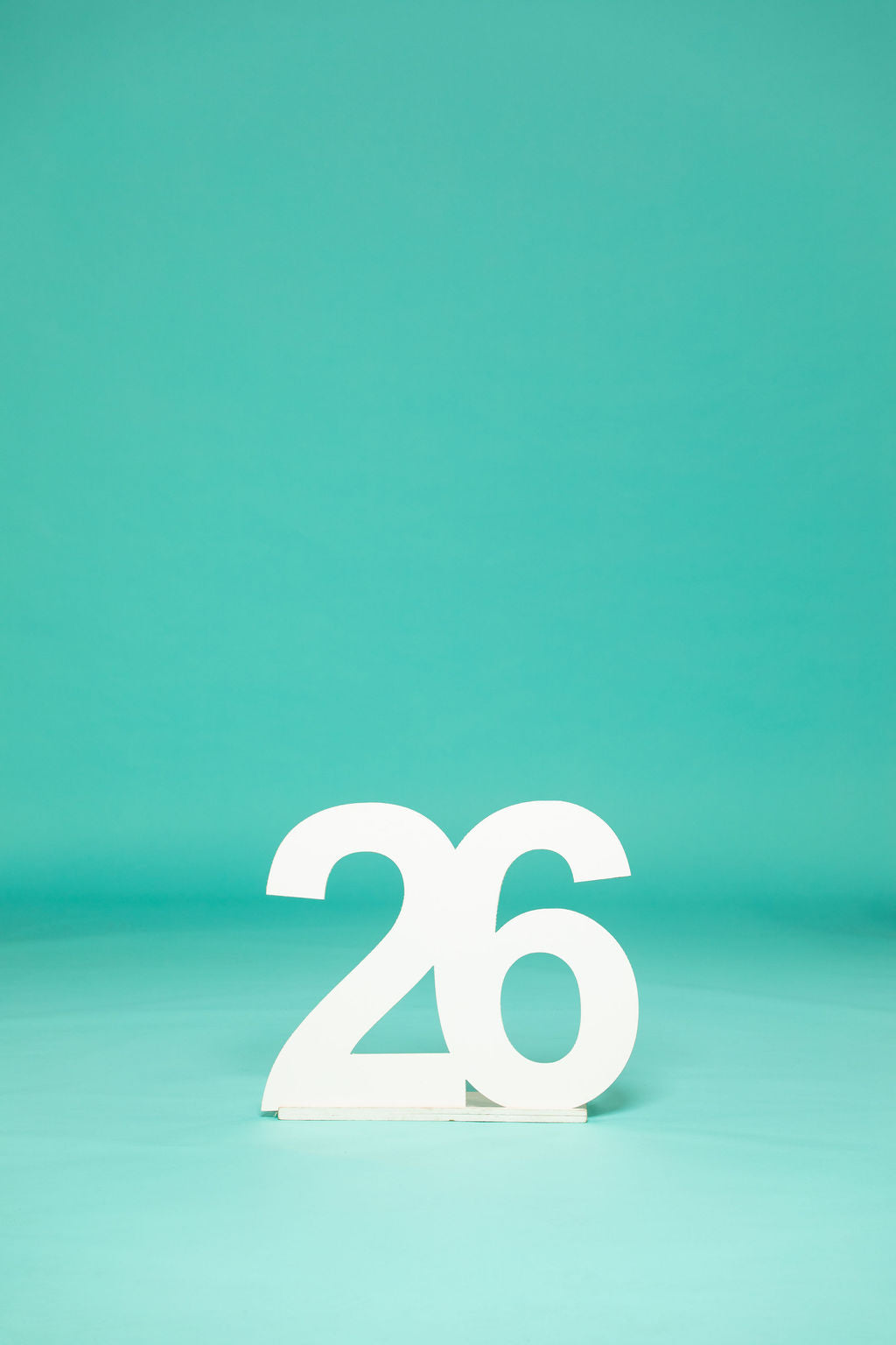 Minimalist shot of a white “26” number prop centered on a teal background, symbolizing a graduating class. Get your 2026 senior props at collageandwood.com.