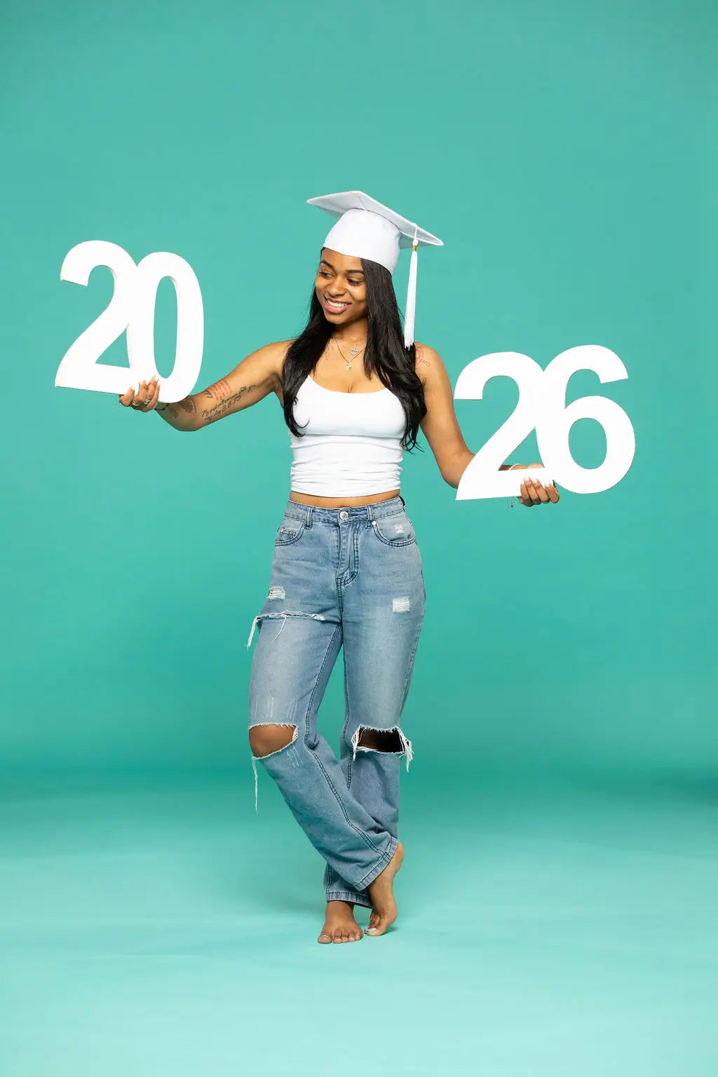 Person in graduation attire holding '2026' photo prop from collageandwood.com on a teal background.