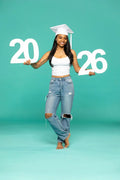 Person in graduation attire holding '2026' photo prop from collageandwood.com on a teal background.