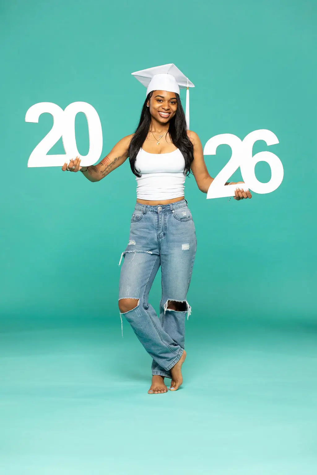 Person in graduation attire holding '2026' photo prop from collageandwood.com on a teal background.