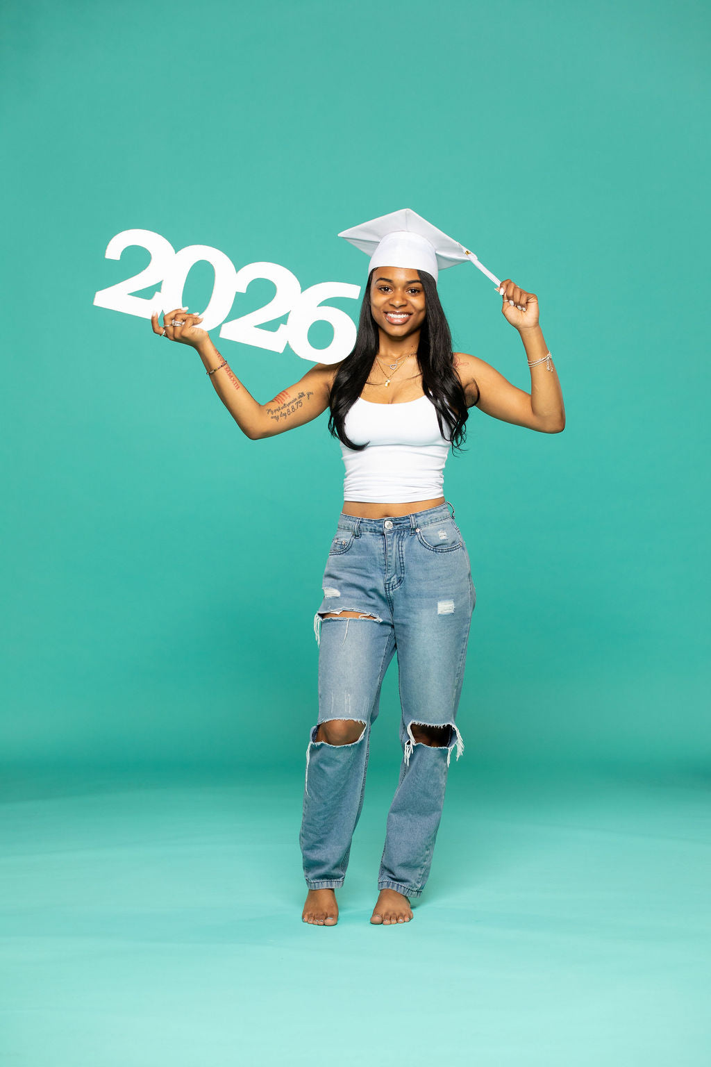 Graduate in white cap holding a bold "2026" sign while smiling confidently in front of a teal backdrop.