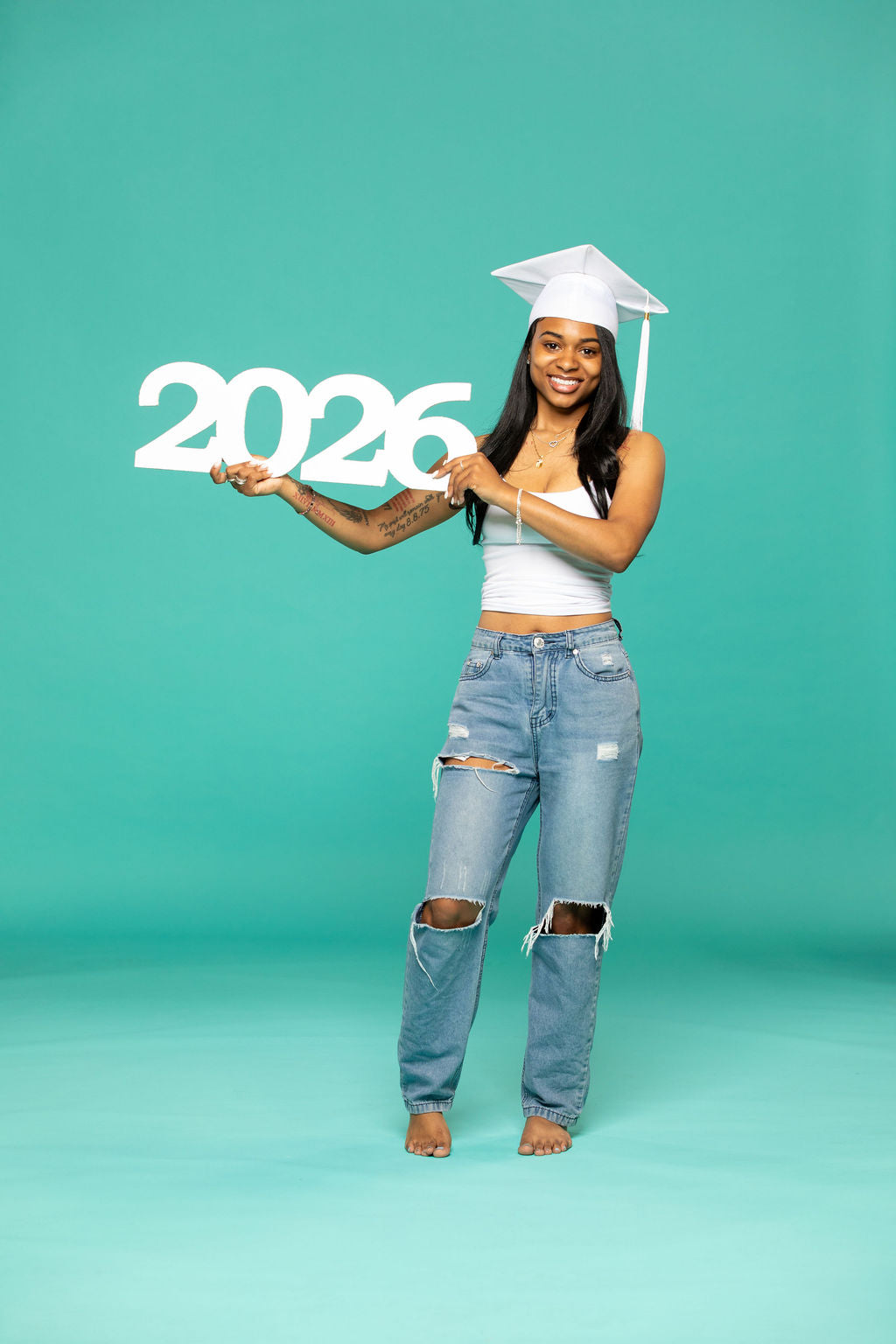 Graduate in white cap holding a bold "2026" sign while smiling confidently in front of a teal backdrop.