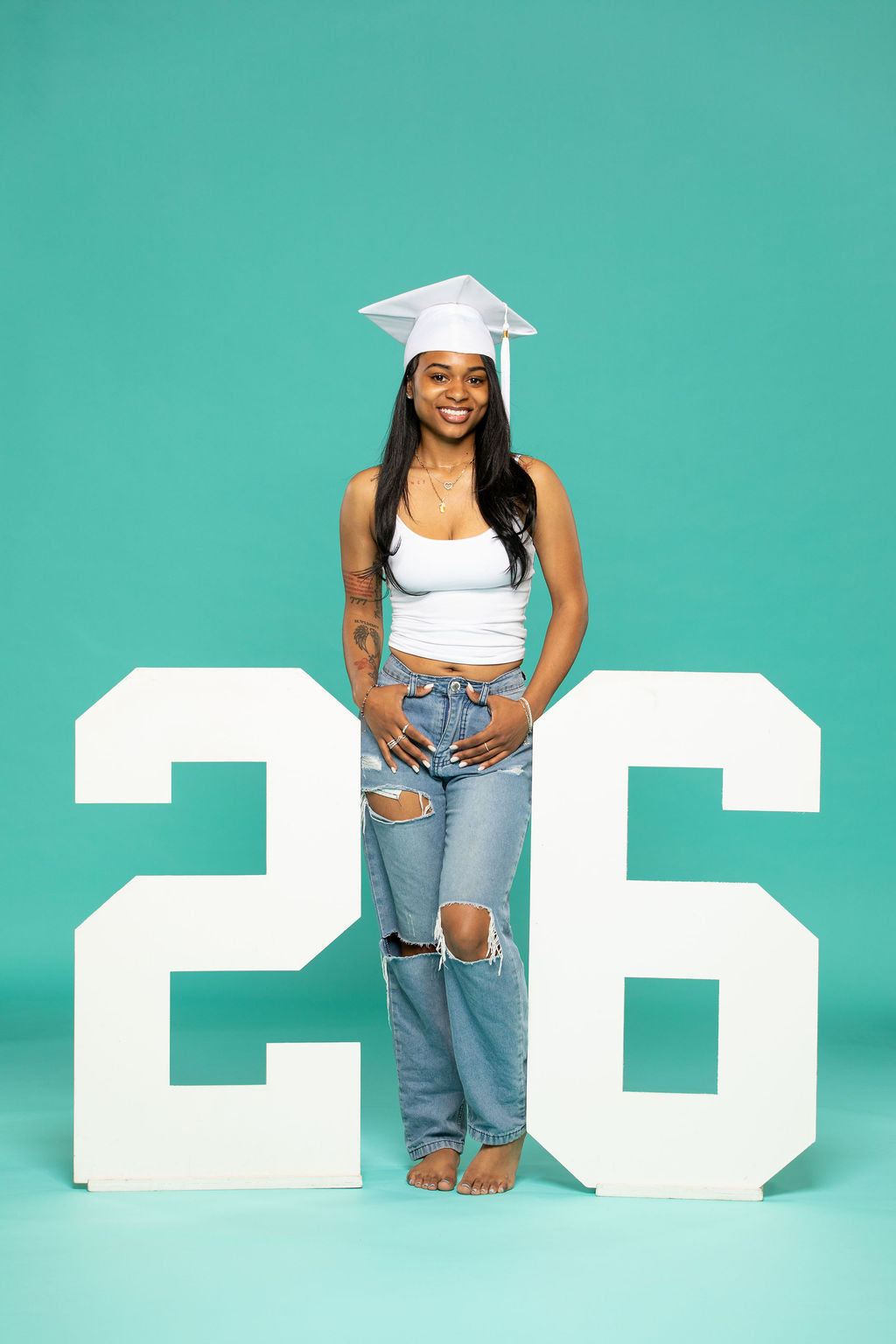 Confident senior girl in white graduation cap and ripped jeans standing barefoot between large "26" props. 2026 senior props available now at collageandwood.com.