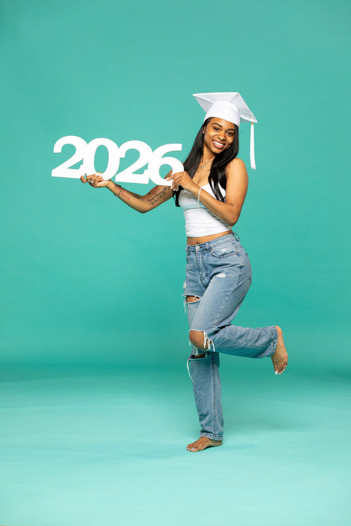 Graduate in white cap holding a bold "2026" sign while smiling confidently in front of a teal backdrop. 