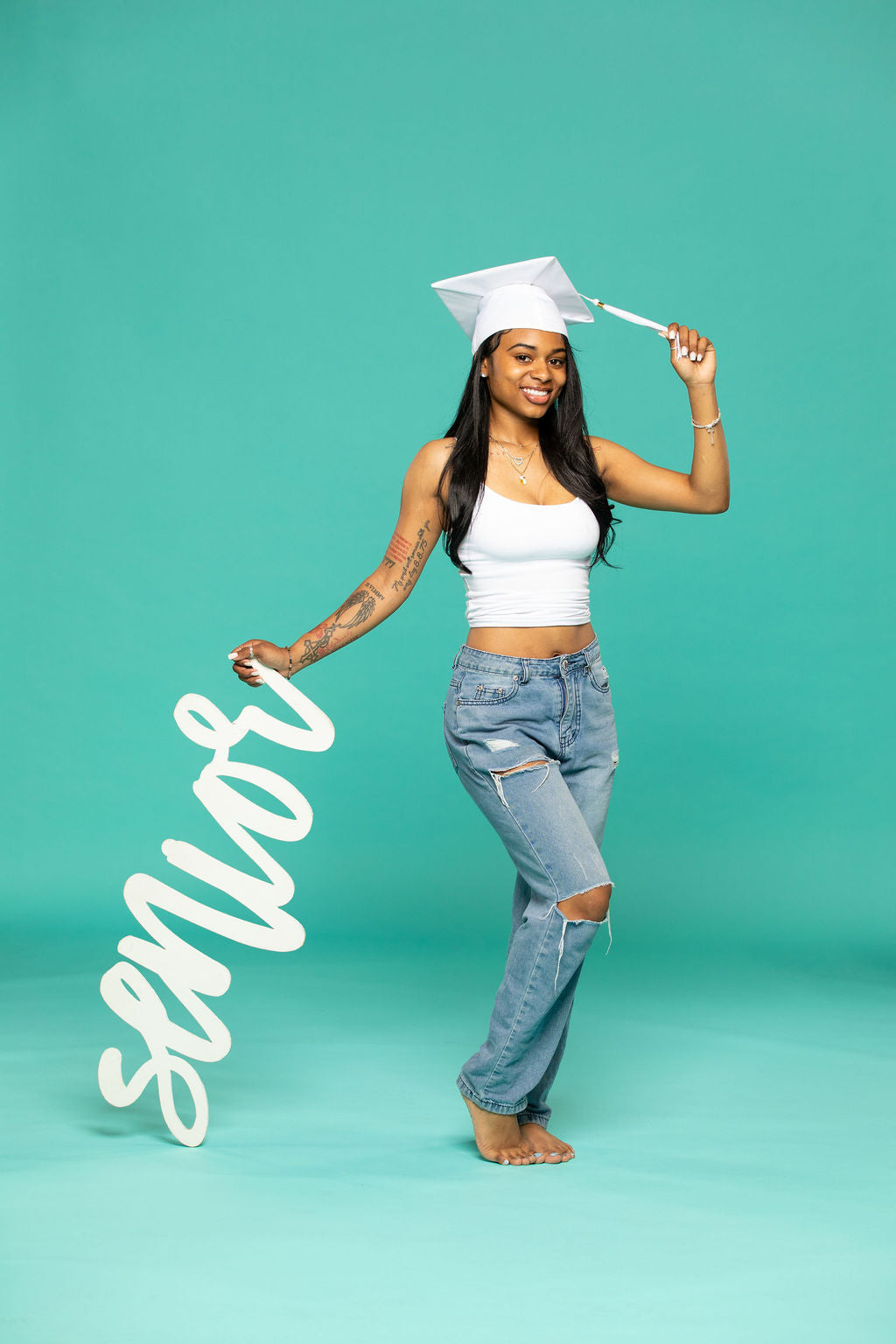 Graduate in white cap holding a flowing “senior” script prop, celebrating with confidence on a teal backdrop. Degree Me props – 2026 senior props available now at collageandwood.com.