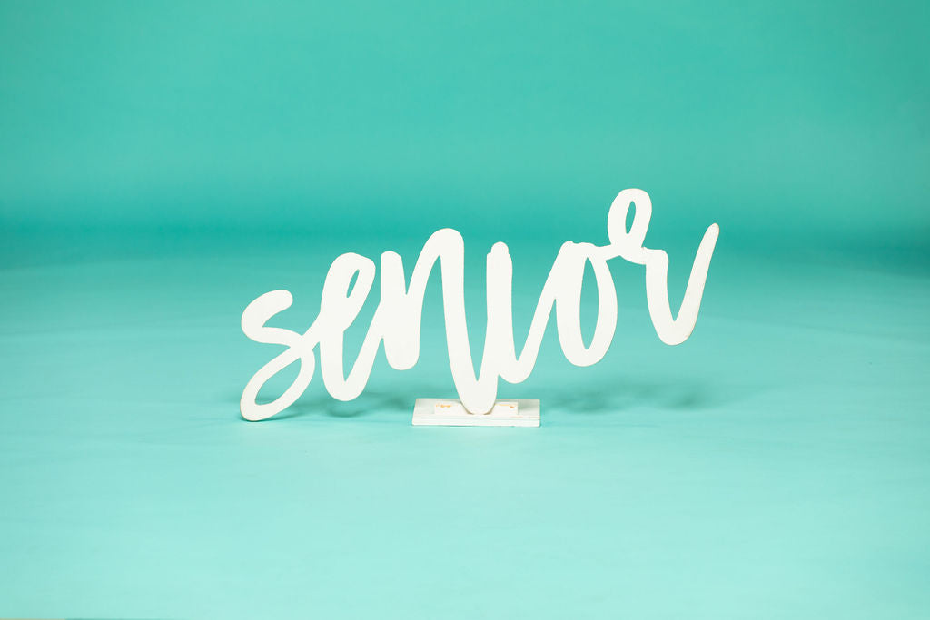 White cursive “senior” sign on a studio floor with teal background, ideal for high school or college graduation photo sessions. 2026 senior props available now at collageandwood.com.