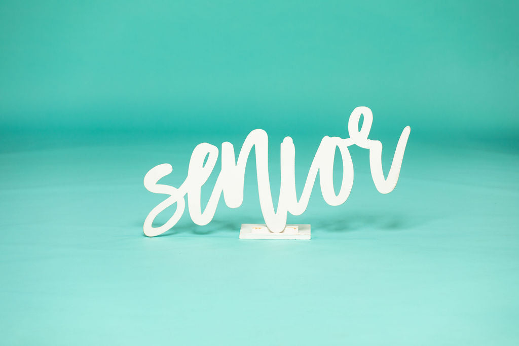 White cursive “senior” sign on a studio floor with teal background, ideal for high school or college graduation photo sessions. 2026 senior props available now at collageandwood.com.