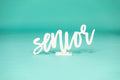 White cursive “senior” sign on a studio floor with teal background, ideal for high school or college graduation photo sessions. 2026 senior props available now at collageandwood.com.