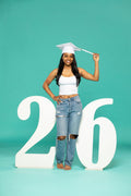 Female graduate in white cap and distressed jeans posing confidently between large script-style “26” numbers, holding her tassel. 2026 senior props available now at collageandwood.com.