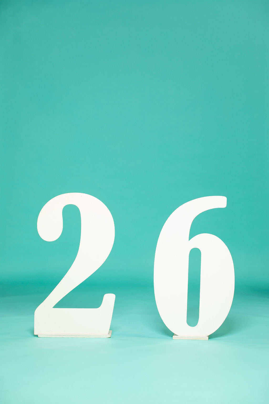 Simple and stylish white number “26” display on a seamless teal backdrop, perfect for 2026 graduation photos. 2026 senior props available now at collageandwood.com.