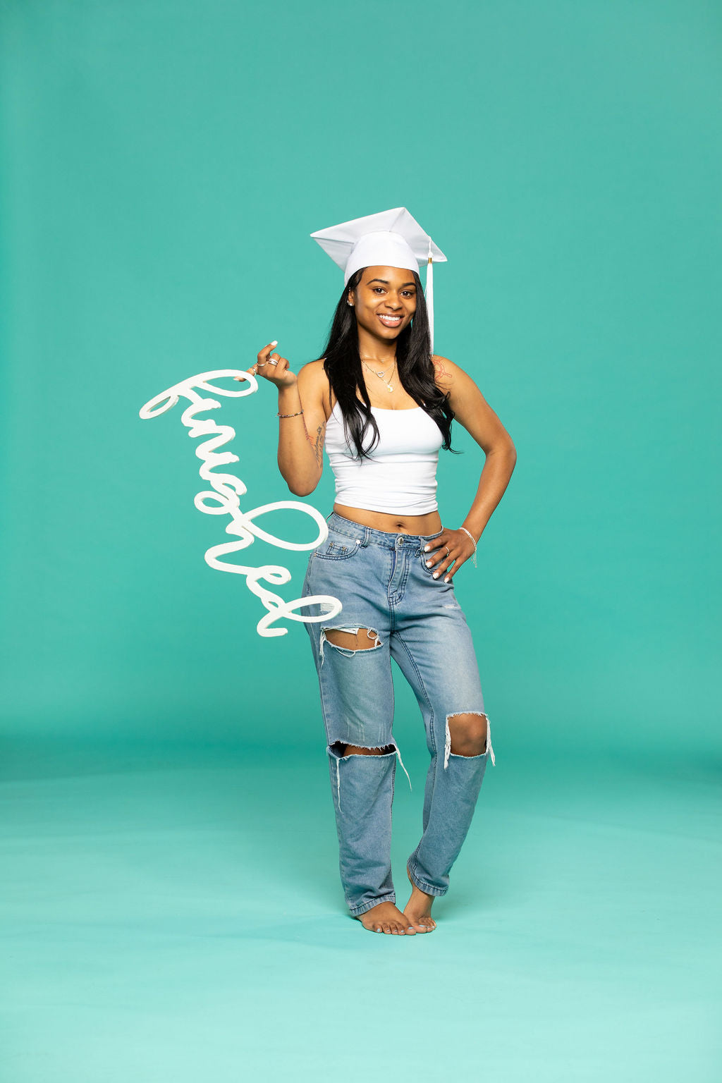 Smiling graduate in a white cap and ripped jeans proudly holding a script-style sign that says “finished” against a teal background. 2026 senior props available now at collageandwood.com.