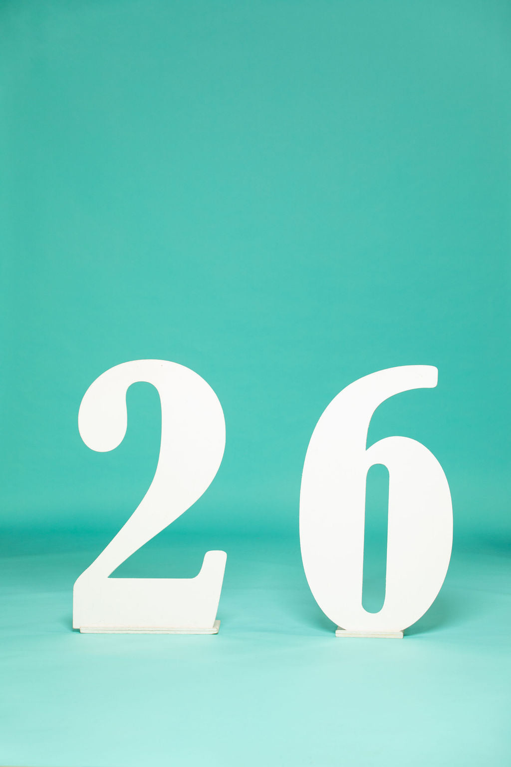 Minimalist setup of bold, curved white numbers “2” and “6” standing upright against a teal background. Ideal for 2026 senior pictures—available at collageandwood.com.
