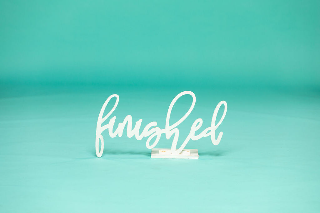 Smiling graduate in a white cap and ripped jeans proudly holding a script-style sign that says “finished” against a teal background. 2026 senior props available now at collageandwood.com.