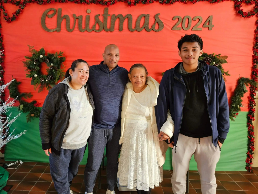 Four family members pose together in front of a festive red and green Christmas 2024 backdrop with holiday wreaths and a white lit tree. A stained glass window and church banner appear in the background, adding to the warm holiday setting.