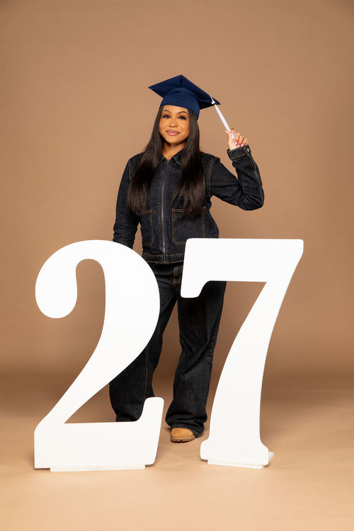 A young woman wearing a dark blue graduation cap and matching jacket and jeans stands behind oversized white numbers '27' on a warm tan studio background. Available at collageandwood.com.