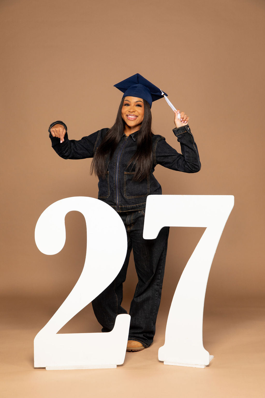 A young woman in a black graduation cap and gown stands behind two large white numbers '27' on a plain tan studio background, holding the tassels of the cap.