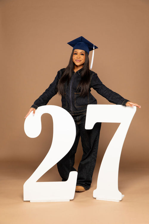 A young woman wearing a dark denim jumpsuit and a navy blue graduation cap stands behind two large white freestanding numbers '27' against a solid tan studio backdrop. Available at collageandwood.com.